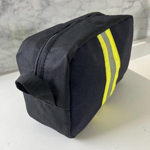 Könnte beinhalten: Schwarze rechteckige Tasche mit Reißverschluss und gewebtem Griff. Die Tasche hat zwei vertikale gelbe Reflexstreifen auf einer Seite. Die Tasche besteht aus einem strapazierfähigen, strukturierten Stoff.