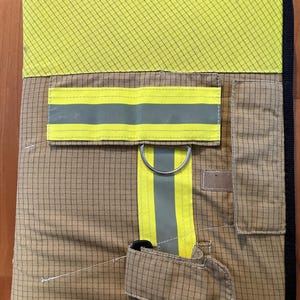 Könnte beinhalten: Beige und gelber Sicherheitsausrüstungsorganisator mit schwarzem Rand. Der Organizer hat ein Gittermuster, reflektierende Streifen und einen Metallring. Eine kleine Tasche und ein Riemen sind ebenfalls sichtbar.