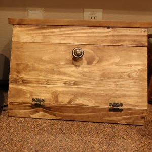 May include: A rectangular wooden bread box with a natural brown wood grain finish. The front features a hinged door with a centered round metal knob and two decorative dark metal hinges at the bottom, allowing the front panel to open outward.