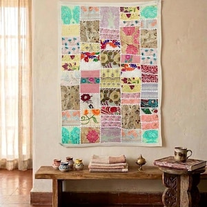 Puede incluir: Un colorido tapiz de pared de patchwork con un borde blanco. El edredón presenta una variedad de telas estampadas florales en tonos de rosa, verde, amarillo y morado. El tapiz se exhibe sobre un banco de madera.