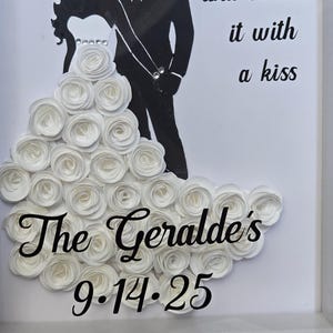 May include: A framed shadow box display featuring a black silhouette of a couple. The figure in the dress is adorned with white paper roses. The text reads: our love story and sealed it with a kiss, The Geralde's, and the date 9.14.25.
