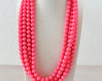 May include: A bright pink beaded necklace with multiple strands. The necklace is displayed on a white mannequin bust. The beads are round and evenly sized, creating a bold and eye-catching accessory.