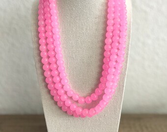 May include: A three-strand pink beaded necklace displayed on a white mannequin. The beads are a uniform size and shape, with a glossy finish. The necklace is draped in a cascading style, showcasing its length and color.