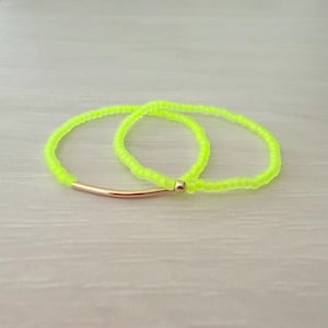May include: Two circular bracelets made of small, neon yellow beads. Each bracelet features a single, smooth, curved gold-toned metal tube accent and a small gold-toned metal bead, creating a bright and simple accessory design.