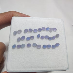 May include: A collection of oval-shaped, pale blue gemstones displayed on a white surface. The gemstones are smooth and polished, with a subtle, translucent quality. The arrangement suggests they are loose stones, possibly for jewelry making.