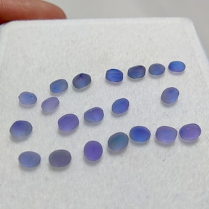 May include: A selection of small, oval gemstones in shades of blue and purple. The stones are displayed on a white surface, with variations in colour and translucency. The gemstones appear to be polished.