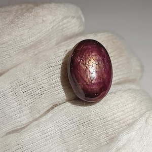 May include: Oval-shaped, polished gemstone with a deep burgundy color and golden internal reflections. The gem is smooth and appears to be held by a white fabric. The stone's surface has a subtle, natural luster, suggesting it is a precious or semi-precious stone.