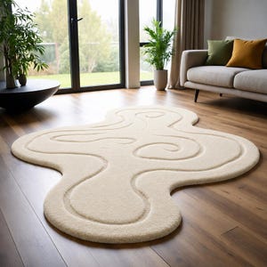 May include: An off-white area rug with an irregular, organic shape and a textured surface featuring carved, swirling line patterns that create a sense of depth. The rug is crafted from a soft, plush material and lies flat on a wooden floor.