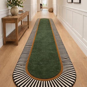 Op de afbeelding: Een lang, ovaal groen tapijt met een bruine rand en een zwart-wit gestreepte rand. Het tapijt ligt in een gang met een lichte houten vloer en witte muren. Het tapijt is ongeveer 300 cm lang.