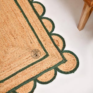 Könnte beinhalten: Ein rechteckiger Teppich aus geflochtener Jute mit einer naturfarbenen Faserstruktur und einem dunkelgrünen, wellenförmigen Rand. Der Teppich hat eine dunkelgrüne rechteckige Innenkante und einen runden Logostempel mit dem Text DAKSH EXPORT.