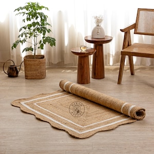 Könnte beinhalten: Ein rechteckiger Jute-Teppich in natürlicher beiger Farbe mit gewelltem Rand. Der Teppich hat ein weißes Bordürenmuster und ist teilweise aufgerollt. Ein kreisförmiges Logo mit dem Text DAKSH EXPORT ist in der Mitte des Teppichs aufgedruckt.