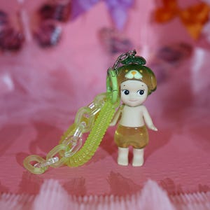 May include: A small figurine of a child-like character with a green hat and yellow shorts, holding a translucent green chain. The figurine has a smiling face and is set against a pink background with blurred bows.