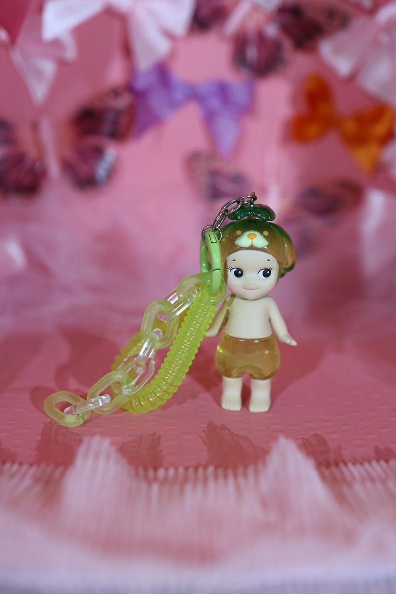 May include: A small figurine of a child-like character with a green hat and yellow shorts, holding a translucent green chain. The figurine has a smiling face and is set against a pink background with blurred bows.