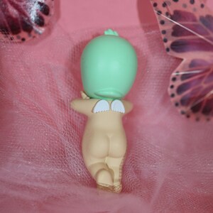 May include: A small figurine of a nude baby with small white wings on its back. The baby has a light green head and is kneeling. The background is pink with a sheer, pink fabric and butterfly decorations.