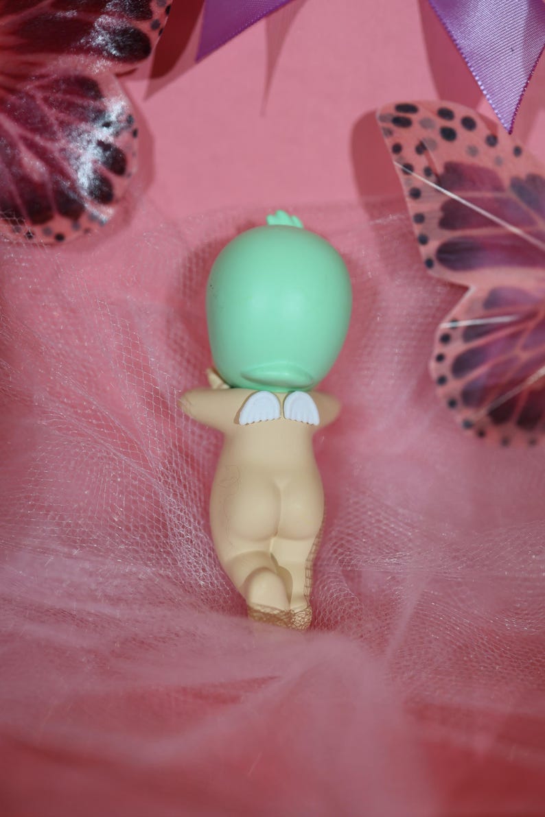 May include: A small figurine of a nude baby with small white wings on its back. The baby has a light green head and is kneeling. The background is pink with a sheer, pink fabric and butterfly decorations.