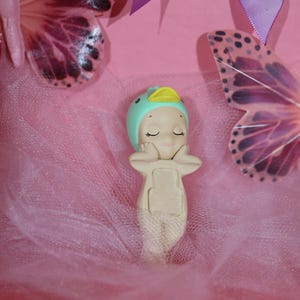 May include: A whimsical figurine of a baby mermaid wearing a teal cap with a yellow duck bill. The figurine is resting on a pink tulle fabric, with two butterfly wings and a purple ribbon in the background. The figurine is beige and has closed eyes.