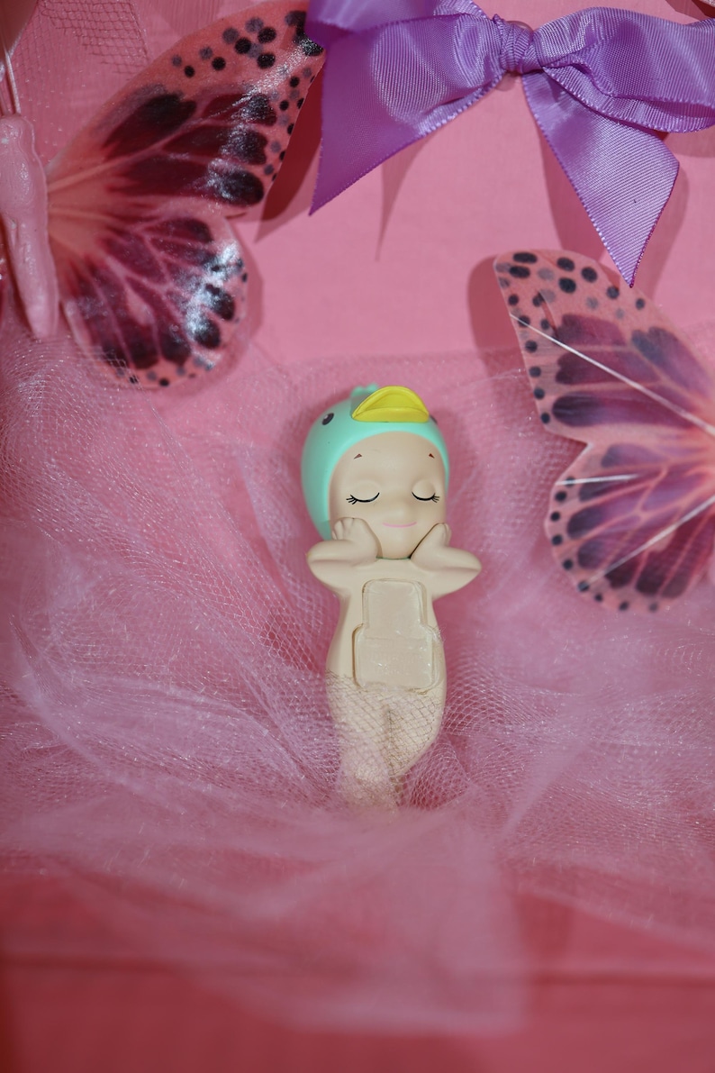 May include: A whimsical figurine of a baby mermaid wearing a teal cap with a yellow duck bill. The figurine is resting on a pink tulle fabric, with two butterfly wings and a purple ribbon in the background. The figurine is beige and has closed eyes.