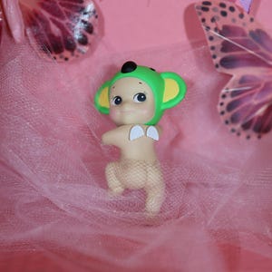 Puede incluir: Una figura caprichosa de una criatura con gorro verde y orejas amarillas, peque&ntilde;as alas blancas y una cubierta similar a una red. La figura est&aacute; sobre un fondo rosa con detalles de mariposas.