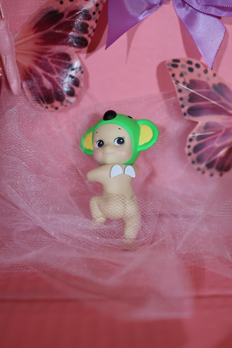 Puede incluir: Una figura caprichosa de una criatura con gorro verde y orejas amarillas, peque&ntilde;as alas blancas y una cubierta similar a una red. La figura est&aacute; sobre un fondo rosa con detalles de mariposas.