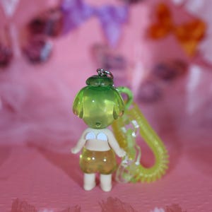 May include: A small, translucent green and yellow figurine of a person wearing a white bikini top and yellow shorts. The figurine has a green head and is attached to a yellow coiled keychain. The background is pink with blurred bows.
