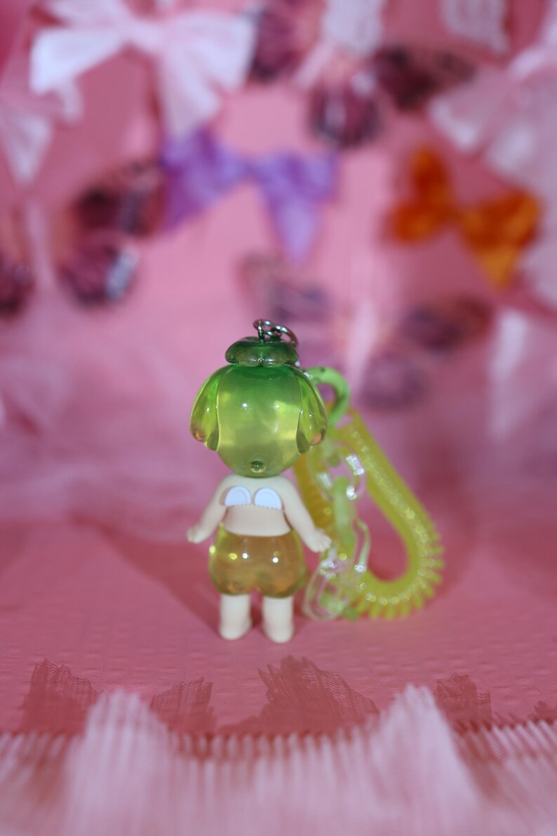 May include: A small, translucent green and yellow figurine of a person wearing a white bikini top and yellow shorts. The figurine has a green head and is attached to a yellow coiled keychain. The background is pink with blurred bows.