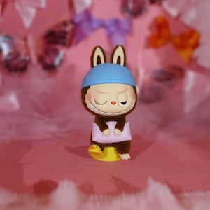 May include: A whimsical figurine of a brown creature with bunny ears, wearing a blue cap, and a wide grin. It's holding a pink pillow and sitting on a yellow duck. The background is a soft pink with decorative bows.
