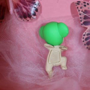 Puede incluir: Una peque&ntilde;a figura verde claro con una cabeza redondeada y un cuerpo beige est&aacute; colocada sobre una tela rosa similar al tul. La figura est&aacute; en posici&oacute;n de rodillas. Alas de mariposa decorativas y una cinta morada en el fondo.