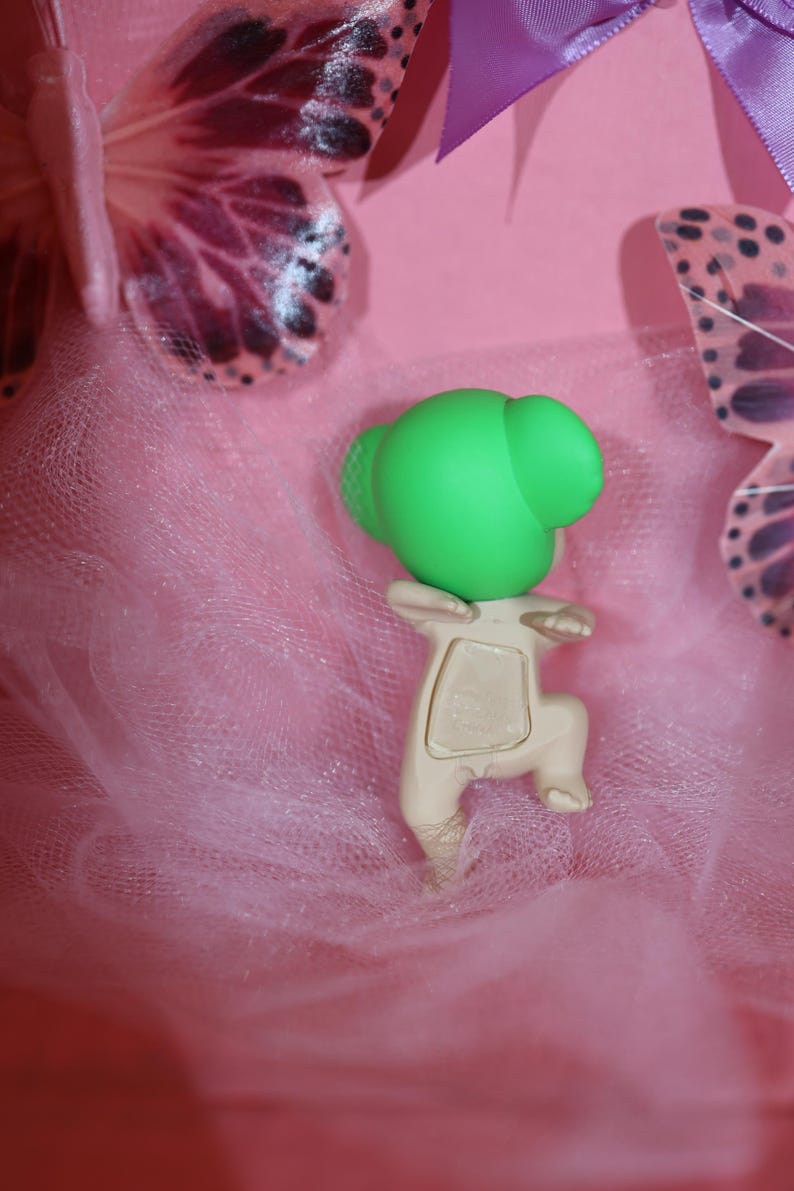 Puede incluir: Una peque&ntilde;a figura verde claro con una cabeza redondeada y un cuerpo beige est&aacute; colocada sobre una tela rosa similar al tul. La figura est&aacute; en posici&oacute;n de rodillas. Alas de mariposa decorativas y una cinta morada en el fondo.