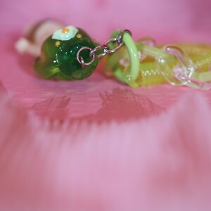 May include: A close-up of a green and yellow charm with a silver chain. The green charm has a flower design with a yellow center. The yellow charm is translucent. The background is a soft pink.