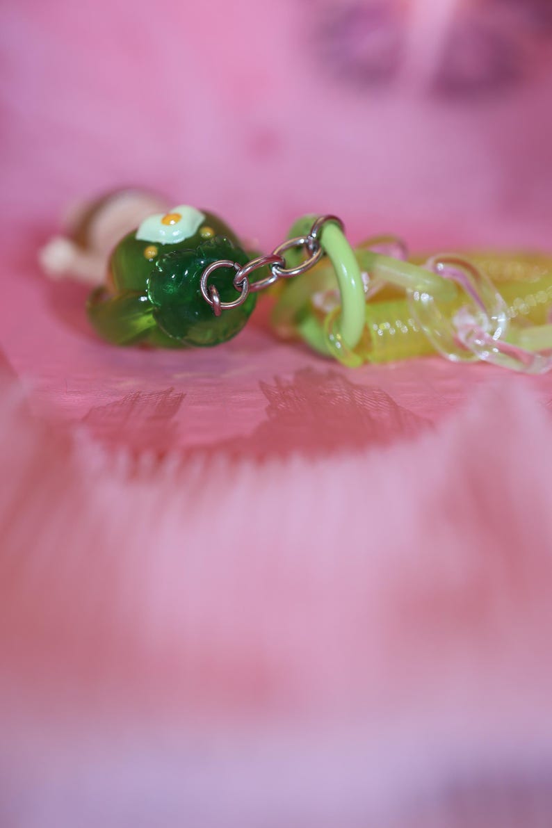 May include: A close-up of a green and yellow charm with a silver chain. The green charm has a flower design with a yellow center. The yellow charm is translucent. The background is a soft pink.