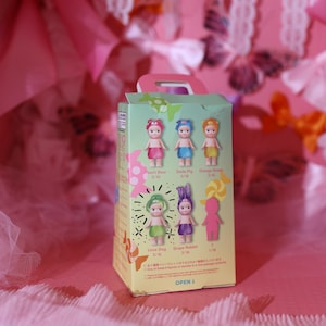 May include: A green and white box featuring six small doll figures with different colored hair and outfits. The box has the words "Peach Bear", "Soda Pig", "Orange Sheep", "Lime Dog", and "Grape Rabbit" printed on it. The box is on a pink surface.