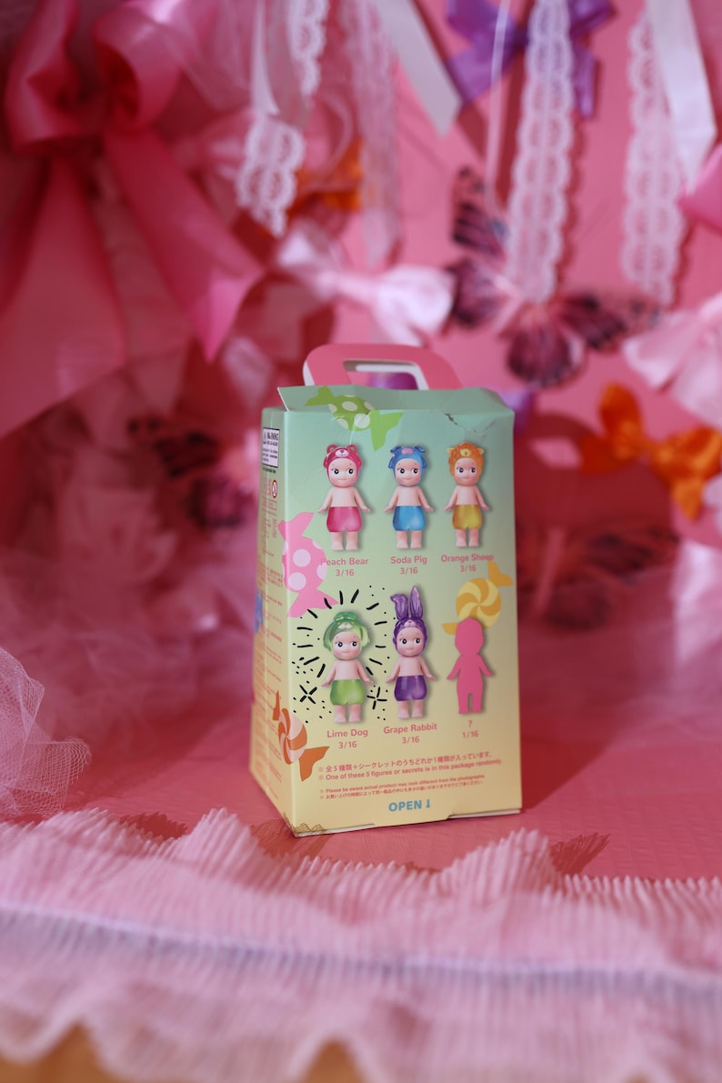 May include: A green and white box featuring six small doll figures with different colored hair and outfits. The box has the words "Peach Bear", "Soda Pig", "Orange Sheep", "Lime Dog", and "Grape Rabbit" printed on it. The box is on a pink surface.