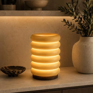May include: A cylindrical table lamp featuring a stack of six smooth, rounded, yellow-toned rings that emit a warm, soft glow. The lamp is supported by a dark, circular wooden base, creating a contrast with the translucent, illuminated body.