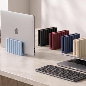 Op de afbeelding: Een collectie verticale laptopstandaards in diverse kleuren, waaronder lichtblauw, zwart, donkerrood, marineblauw, crème, grijs en olijfgroen. Elke standaard heeft een geribbeld, U-vormig ontwerp om een laptop rechtop op een bureau te houden.