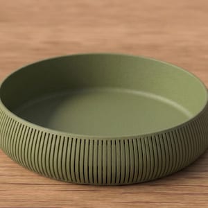 May include: An olive green, round bowl with a textured, ribbed exterior. The bowl sits on a wooden surface. The bowl's design features vertical lines around the sides, adding a decorative element to the piece.