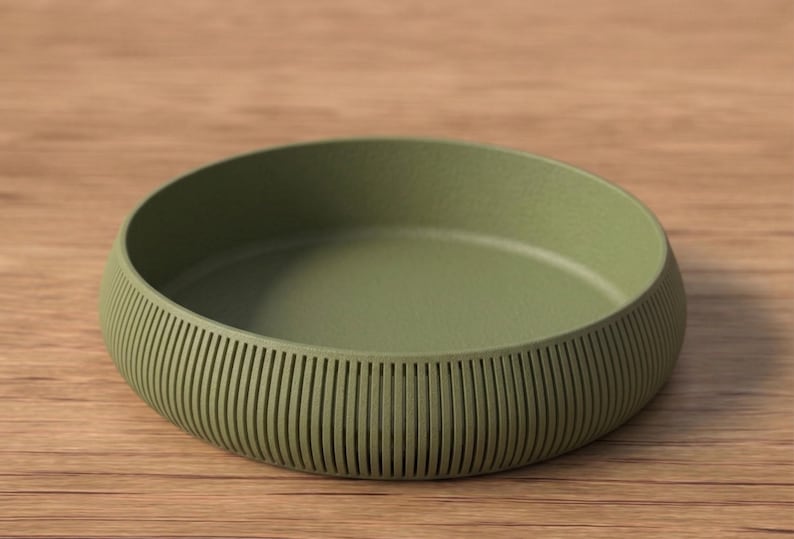 May include: An olive green, round bowl with a textured, ribbed exterior. The bowl sits on a wooden surface. The bowl's design features vertical lines around the sides, adding a decorative element to the piece.
