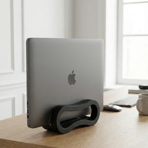 Op de afbeelding: Een grijze laptop met het Apple-logo wordt rechtop gehouden door een zwarte, gebogen standaard. De standaard staat op een lichtbruin houten bureau. Een toetsenbord, muis en mok zijn zichtbaar op de achtergrond.