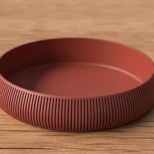May include: A terracotta-colored, round bowl with a low profile. The exterior features vertical, grooved detailing, while the interior is smooth. The bowl sits on a wooden surface.