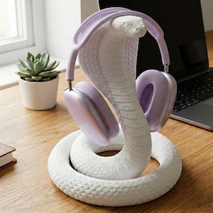 Peut inclure: Un support de casque en forme de cobra blanc avec une surface texturée, tenant une paire de casques lavande. Le support est sur un bureau en bois, avec un ordinateur portable, une tasse et une plante succulente en arrière-plan.