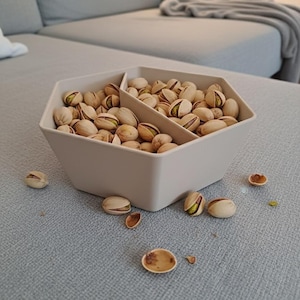 May include: A beige, hexagonal serving bowl filled with pistachios. The bowl is divided into two sections. Several pistachios and shells are scattered around the bowl on a gray surface. The pistachios have a light tan shell and a green interior.