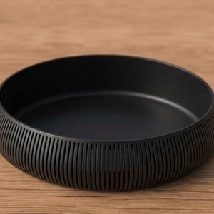 May include: A matte black bowl with a low profile and a textured, ribbed exterior. The bowl sits on a wooden surface. The bowl's design is simple and modern, suitable for serving or display.
