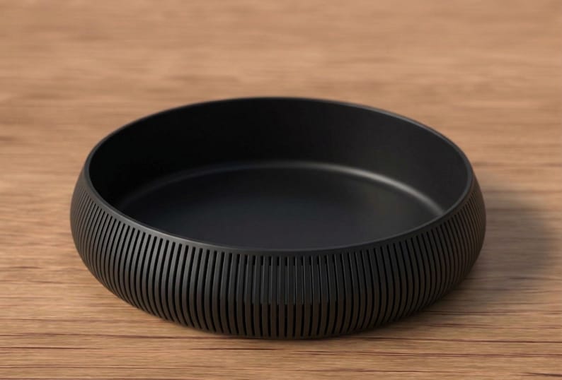 May include: A matte black bowl with a low profile and a textured, ribbed exterior. The bowl sits on a wooden surface. The bowl's design is simple and modern, suitable for serving or display.