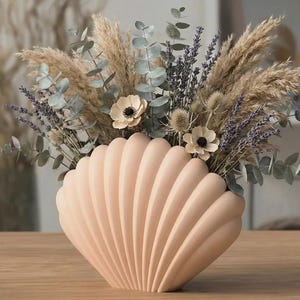 Könnte beinhalten: Eine pfirsichfarbene, muschelförmige Vase gefüllt mit einem Trockenblumenarrangement. Das Arrangement umfasst Pampasgras, Eukalyptus, Lavendel und Anemonenblüten. Die Vase steht auf einer Holzoberfläche und schafft eine natürliche und elegante Präsentation.