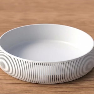 May include: A white, round bowl with a ribbed exterior design. The bowl has a shallow depth and sits on a wooden surface. The bowl's design features vertical lines around the exterior, creating a textured appearance. The bowl is likely used for serving or display.