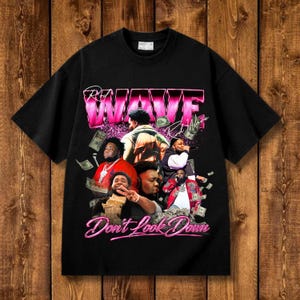 May include: A black short-sleeve t-shirt featuring a graphic print of the artist Rod Wave. The design includes the text "ROD WAVE" in large pink letters, "Don't Look Down" in pink script, and multiple images of the artist with floating US dollar bills.