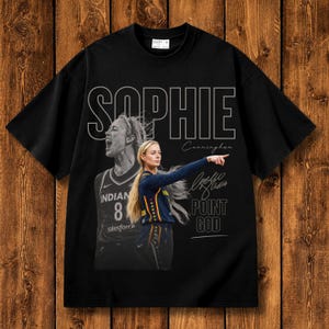 Puede incluir: Una camiseta negra de manga corta con un estampado gráfico de la jugadora de baloncesto Sophie Cunningham. El diseño incluye el nombre SOPHIE en letras blancas grandes, una imagen en escala de grises, una imagen a color y el texto POINT GOD.