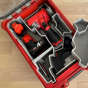 May include: Red toolbox with gray compartments containing power tools, including a drill, batteries, and wrenches. The toolbox is open, revealing the organized contents. A hand is holding a gray compartment.