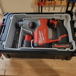 May include: A red and black Milwaukee FUEL hammer drill kit in a black hard case. The case is open, revealing the drill, battery, and drill bits. The drill has a black handle and a red body. The case is on a wooden surface.
