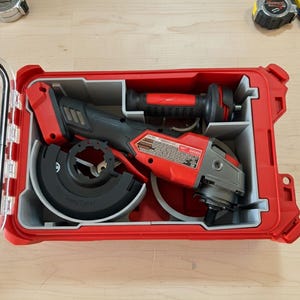 Puede incluir: Una amoladora angular roja y negra con protector y mango negros, guardada en una caja roja con compartimentos grises. La caja está abierta, revelando la herramienta y los accesorios. Un flexómetro y un destornillador son visibles cerca.