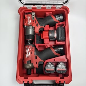 Pode incluir: Um kit de ferramentas elétricas Milwaukee vermelho com tampa transparente. O kit contém duas ferramentas elétricas vermelhas e pretas, duas baterias pretas e um carregador. As ferramentas estão dispostas em uma caixa de plástico vermelha.